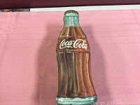 Photo of Coca Cola bottle shaped metal tin laying on pink surface showing front branding and embossed bottle design.