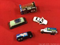 Five die cast metal cars displayed on red background including Hot Wheels black truck, white police car, blue and white delivery truck, mint green convertible, and boxed Maisto Power Truck Racer
