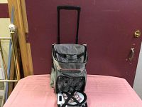 Gray rolling cooler cart with telescoping handle shown upright on table with picnic set resting in front.