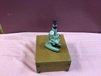 Gray metal microscope standing on closed wooden box with metal handle.
