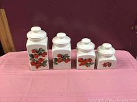 Full front view showing all four ceramic canisters arranged by descending height on a pink cloth, against a red background.