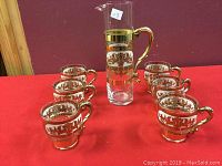 Full set of one glass pitcher and six matching glasses with decorative gold trim and pattern on a red cloth background.