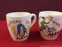 Two vintage ceramic coffee mugs featuring cartoon Ma and Pa characters with humorous phrases on the front, placed on a red surface against a dark background.