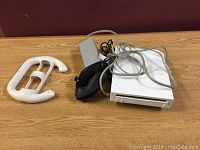 Full view of Nintendo Wii console with Wii remotes, power adapter and Wii wheel accessory on wood surface.