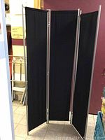 Front angle view of the black cloth and metal dressing screen showing its three panels fully opened in an indoor setting.