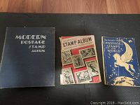 Three stamp albums arranged side by side on a wooden floor, titles visible as 'Modern Postage Stamp Album,' 'The Adventuresome Stamp Album,' and 'The Adventuresome Album Postage Stamps of the World'.