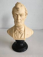 Front and top front angled view of the beige Abraham Lincoln bust on a black base, showing face, hair, beard, bow tie, and the name LINCOLN inscribed.