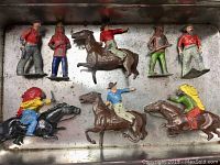 Top-down view of eight hand-painted lead cowboy and Indian figures, some mounted on horses, showing varying postures and colors in a metal tray.