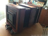Large antique wooden camera case with expandable bellows shown from a front side angle on a wooden table indoors.