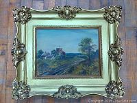 Full view of antique framed oil painting showing rural scene with a road, houses, and a church tower, in an ornate gold frame.