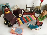 Overall view of the ten vintage toys including tin toys, wooden xylophone, animal figurines, binoculars in box, and toy sewing machine.