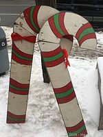 Two large wooden candy cane decorations leaning against a tree outdoors in a snowy setting, showing striped red and green paint with red bows.