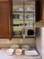 Open kitchen cabinet showing shelves stocked with various white ceramic plates, bowls, and cups with a dark rim detail characteristic of Dansk bistro set.