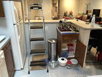 Kitchen corner with stainless steel trash can, step stool, Pyrex bowls, baking pans and some plastic containers shown on floor and cabinet under counter shows stacked plastic food storage containers with lids and two squeeze bottles.
