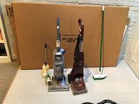 Wide view showing all items in the lot including Bissell QuickSteamer, Hoover FloorMate, unopened yellow duster, green push broom, and two cleaning fluid bottles in front.