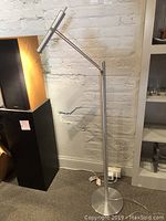 Full-vertical view of the entire stainless steel floor lamp showing the base, adjustable arm, and lamp head against a white brick wall background.
