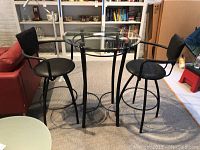 Full view of glass top cafe table with two black swivel stools beside a red sofa in a room with shelves and carpet.