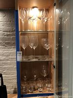 Wooden cabinet with upper shelves holding multiple tall wine glasses with long stems and slightly flared bowl shapes, accompanied by smaller glasses below.