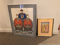 Two framed artworks on carpet floor: larger Korean wedding portrait print on the left, smaller Indian fabric painting on the right.