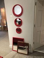 Wide photo showing all five mirrors displayed on and below the wall: three round red metal framed mirrors vertically aligned on the wall, rectangular red mirror and square wood mirror leaning at bottom.