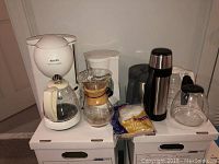 Full view of two coffee makers, Chemex, extra carafe, travel mug, and coffee filters on white boxes against wall