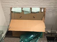 Photo showing the glass tabletop leaning on a wall partially covered with cardboard and protective wrapping, highlighting its size and packaging.
