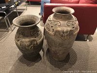 Photo showing two large clay floor vases with elephant heads and embossed swirl patterns, placed on carpeted floor in living room.