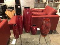 Full view of all 5 vases placed on a glass table in a living room setting, showing four red ceramic and one glass vase