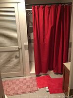 Photo of a bathroom with red Nautica fabric shower curtain hanging on metal hooks, and two rugs on the floor: a plain red bath mat and a red and white woven throw rug.