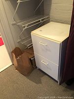 White MDF 2-drawer file cabinet with two front-facing pull handles, placed on carpeted floor near metal wire shelving and cardboard box.