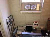 Wide angle photo showing all items in the lot placed on and around a table beneath a window. Items include a walker, binding machine, shredder, heater, blood pressure monitor, phone, and toilet seat.