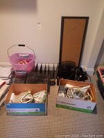 Overview photo showing two numbered cardboard boxes filled with assorted tangled cords (power, extension, computer, AV cords), a pink plastic bucket, black metal mesh letter trays, a small cork bulletin board, and a black desk lamp.