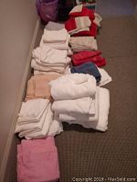 Overview image showing folded sheets in white, taupe, red, and pink along with some towels on a carpeted floor.