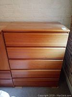 Full front view of the clean-lined mid-century modern wooden chest of drawers against a brick wall, showing five horizontal drawers closed.