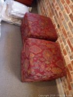Two cube ottomans with red multicolored geometric patterned fabric upholstery placed on carpet floor against brick wall.