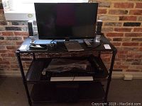 Samsung 24-inch TV/computer monitor on a black stand with pair of Creative speakers on each side, four keyboards below the TV on shelving and subwoofer under the bottom shelf.