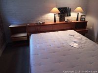 Two mid-century modern wood side tables placed on either side of a bed with a headboard featuring multiple drawers and a shelf area, lit by wall lamps.