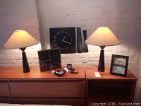 Wide shot showing both clocks (one square black and one square with silver frame), two black table lamps with beige shades, two black vases, travel clocks, Casio watch and picture frame