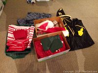 Box with folded red and white linens, red kitchen towels, oven mitts, and checked napkins, plus Christmas-themed items and a black wizard costume with hat.