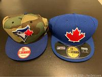 Front view of two Blue Jays baseball hats: the left one is camo with Blue Jays logo, the right one is blue with red maple leaf.