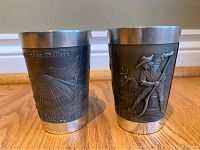 Photo of both Degussa pewter cups showing detailed raised relief artwork of outdoor and work scenes on wooden floor with white baseboard.