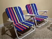 Two folding beach chairs side by side showing the colorful vertical striped upholstery, white metal frame, and white armrests.