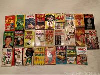 Top-down photo of all 25 vintage MAD Magazine books laid out flat on a white surface showing colorful covers and variety of titles.
