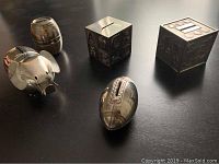 Five metal piggy banks displayed on a table: an elephant, a football, a barrel, and two cubes with embossed designs and coin slots on top.