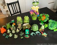 Multiple frog figures and accessories arranged on a dark table showing the variety of the collection, including large and small figurines, planters, and canisters.