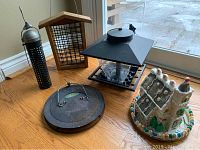 Overview of five different bird feeders including tube feeder, wooden framed feeder, lantern style feeder, mesh dome feeder, and ceramic decorative house feeder, all placed on a wooden floor near glass door