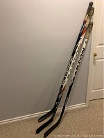 Two right-handed hockey sticks leaning against wall, taped handles and blades, Easton and Bauer brands visible