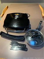 George Foreman grill, egg poacher pan with lid, two black spatulas on table.