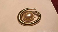 Front view of gold-tone swirled abstract brooch with central oval faux pearl on beige surface