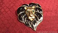 Close-up image of a lion head brooch on a textured red background showing black enamel mane, gold-tone face, and green crystal eyes.